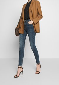 Brun blazer, svart skjorta, blå smala jeans, svarta högklackade sandaler med remmar och en mönstrad handväska.