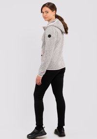 Grauer Hoodie aus strukturiertem Stoff, pinken Akzenten und einem schwarzen Logo-Patch. Kombiniert mit schwarzen Leggings und schwarzen Sportschuhen.