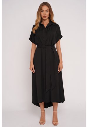 Kobieta w czarnej, midi sukience z krótkim rękawem, kołnierzykiem, wiązanej w talii, oraz beżowych sandałach na wysokim obcasie, stojąca na jednolitym tle.