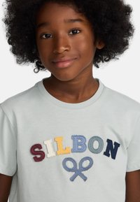Camiseta de algodón azul claro con letras bordadas de colores "SILBON" y un diseño de tijeras azules. Cuello redondo, mangas cortas.
