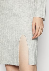 Vestito grigio a maglia a coste con uno spacco laterale. Primo piano del tessuto che mostra una consistenza morbida e un design aderente. Mano posata sul vestito.