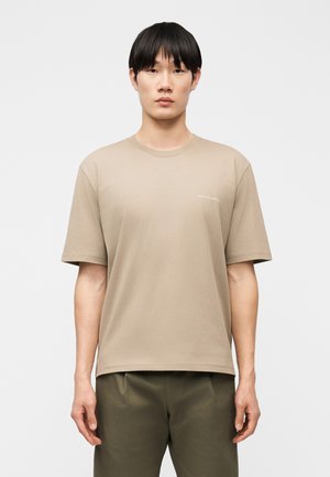 Giovane uomo con capelli neri lisci che indossa una maglietta beige a maniche corte e pantaloni verde oliva su uno sfondo chiaro uniforme.