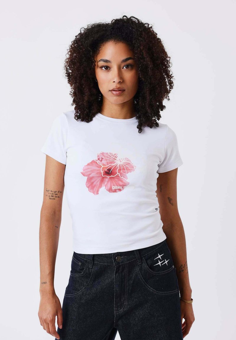 Femme aux cheveux bouclés portant un t-shirt blanc avec un motif de fleur rose et un pantalon noir avec des symboles d'avion blancs cousus.