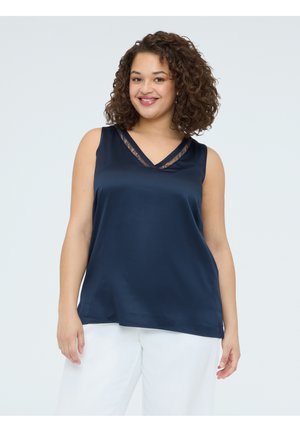 Lächelnde Frau mit lockigem Haar, die ein ärmelloses dunkelblaues Oberteil mit Spitzenbesatz und weiße Hose trägt, vor einem hellen Hintergrund stehend.