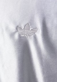 Detailní záběr na bílou texturovanou tkaninu s vyšitou logem Adidas trefoil ve stejné bílé nitce.