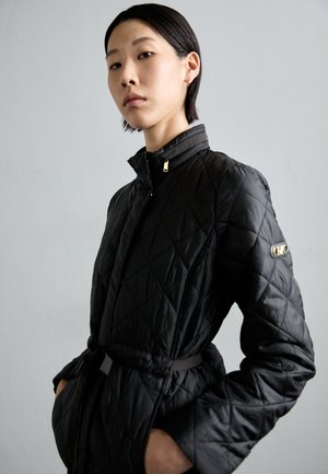 Giacca nera trapuntata con colletto alto, zip frontale e vita con cintura. Presenta un accento con logo sulla manica sinistra e una texture liscia.