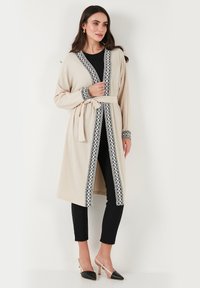 Beige long cardigan with black embroidered trim, open front, tie belt, and long sleeves over a black top and slim pants.