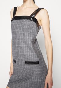 Femme portant une robe grise à carreaux sans manches avec des bretelles noires brillantes et une bordure assortie sur les poches avant.