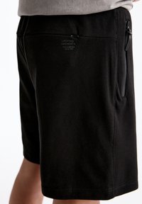 Shorts noirs en tissu texturé, avec une poche zippée sur le côté et une étiquette logo à l'arrière. Le design est minimaliste et fonctionnel.