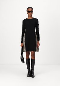 Schwarzes langärmeliges Kleid mit animalischen Druckseitenteilen, runder Halslinie und kontrastierenden weißen Streifen an den Ärmeln; kombiniert mit kniehohen schwarzen Stiefeln.