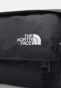 Borsa messenger nera con finitura lucida, dotata di un logo bianco, tracolla regolabile e una patta superiore con chiusura a scatto.