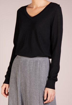 Kvinde iført en sort V-hals langærmet sweater kombineret med et højtaljet, gråt plisseret skørt, stående mod en neutral baggrund.