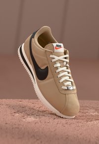 Rudos sportiniai kedai su juodu swoosh, baltais raišteliais ir sidabro detalėmis. Apvalus pirmas pirštas ir tekstūruotas paviršius, įmontuoti ant balto guminio pado.