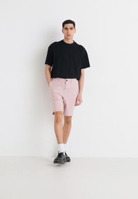 T-shirt noire à manches courtes, shorts roses clairs avec fermeture à bouton, et chaussures noires. Le modèle se tient devant un fond blanc uni.