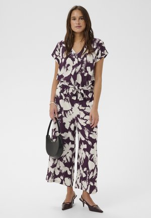 Femme portant une combinaison à fleurs violettes et blanches, à manches courtes et jambes larges, tenant un sac à main noir et portant des talons foncés.