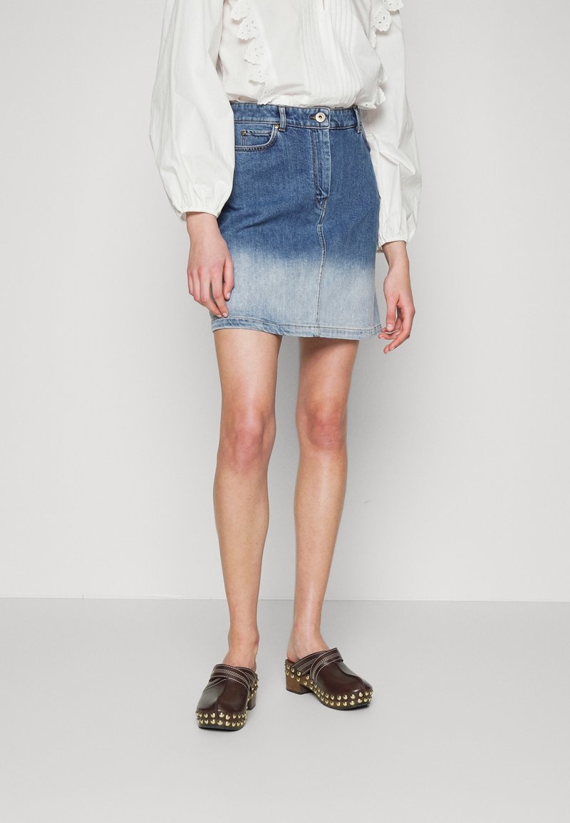 Femme portant une jupe en jean dégradé bleu, un chemisier blanc à manches longues avec des volants, et des sabots marron foncé cloutés, debout devant un fond uni.