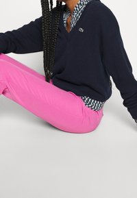 Un pull col en V bleu marine porté par-dessus une chemise à col à motifs, associé à un pantalon rose vif. La tenue présente une finition texturée et un logo brodé.