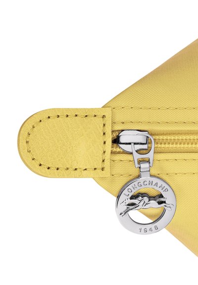 Yellow bag corner with visible zipper and silver round Longchamp 1948 logo charm attached to the zipper pull tab.