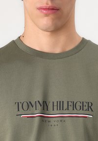 T-shirt de algodão verde-oliva com um logo preto "TOMMY HILFIGER" e riscas vermelha e branca ao centro. Design padrão de decote redondo.
