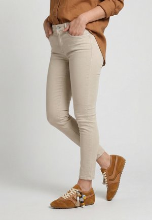 Persoon draagt beige skinny broek, bruine casual schoenen met decoratieve kwastjes en een bruine blouse met lange mouwen, staand met handen in de zakken.