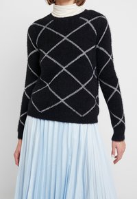 Zwarte pluizige trui met wit ruitpatroon, ronde halslijn en lange mouwen, gecombineerd met een lichtblauwe geplooide rok.