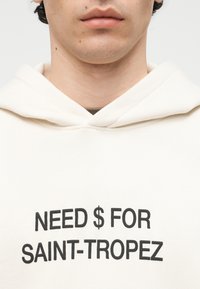 Gros plan sur une personne portant un sweat à capuche blanc avec un texte noir centré sur la poitrine indiquant « NEED $ FOR SAINT-TROPEZ ».