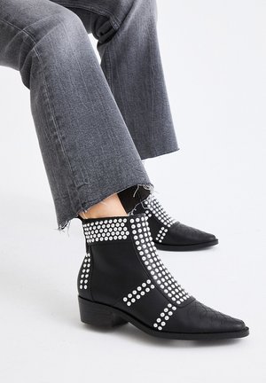 Botas negras hasta el tobillo con punta afilada, que presentan una superficie texturizada, acentos de tachuelas y un tacón cuadrado bajo. Combinadas con jeans grises deshilachados.