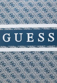 Niebieski i biały materiał z powtarzającym się wzorem logo, z wyraźnym logo "GUESS" wytłoczonym dużymi białymi literami, umieszczonym poniżej na środku.