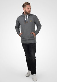 Sudadera gris con capucha alta, cordones blancos y detalles en tono madera. Combinada con jeans negros desgastados y zapatillas blancas.