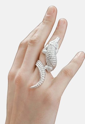Anillo de cocodrilo plateado con escamas texturizadas, cola curvada y detalles realistas, llevado en un dedo con un tono de piel natural.