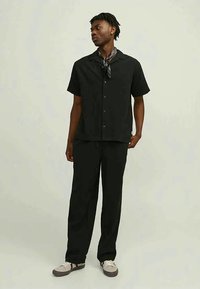 Jovem vestido com uma camisa preta de manga curta e calças, usando um lenço de pescoço estampado e ténis claros, de pé contra um fundo simples.