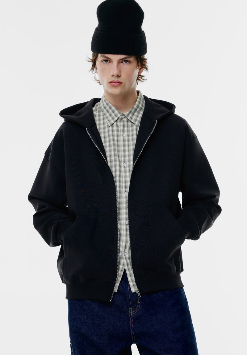 Felpa nera con cappuccio e zip anteriore, indossata sopra una camicia a quadri verde chiaro. Il modello indossa anche un cappellino nero e jeans blu.