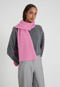 Sciarpa di lana rosa drappeggiata su un maglione grigio lavorato a trecce, con una texture morbida e una forma rettangolare, che completa un paio di pantaloni grigio chiaro.