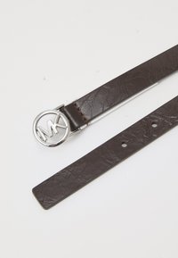 Cintura in pelle marrone con una fibbia metallica circolare argento con le iniziali "MK", texture liscia e diversi fori di regolazione per la misura.
