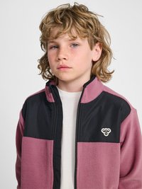 Blond kind met krullend haar en sproeten draagt een roze-zwart fleecejack met rits over een wit shirt, kijkt recht vooruit met een neutrale gezichtsuitdrukking.