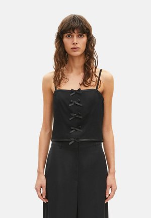 Kvinde med bølget brunt hår iført en sort ærmeløs top dekoreret med fem små sorte sløjfer ned langs fronten.