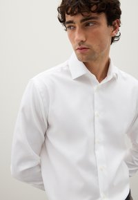 Giovane uomo con capelli ricci scuri che guarda a destra, indossando una camicia bianca con bottoni su uno sfondo chiaro e uniforme.