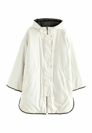 Femme portant un manteau à capuche beige clair avec boutons et fermeture éclair, mains dans les poches, debout à l'extérieur près d'une clôture métallique noire.