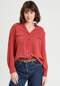 Chemise rouge à boutons avec manches longues, deux poches poitrine et décolleté en V, portée par-dessus un jean taille haute bleu avec une ceinture à imprimé léopard.