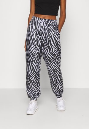 Personne portant un crop top noir et un pantalon jogging imprimé zèbre noir et blanc avec des baskets blanches, debout devant un fond uni.
