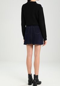 Schwarzer Strickpullover, dunkelblauer Jeansrock, Stiefeletten. Der Pullover hat einen hohen Kragen, der Rock verfügt über zwei Gesäßtaschen und Saumdetails.