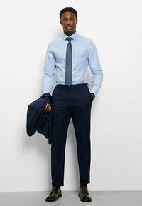 Hellblaues Hemd mit spitzem Kragen, marineblauer Krawatte und dunkelblauen Hosen. Ein marineblaues Sakko über einem Arm gelegt und polierte braune Schuhe.