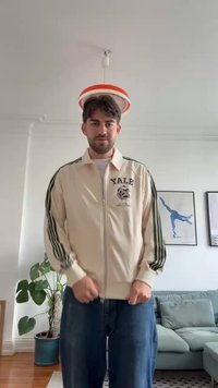 Kremfarget zip-up jakke med grønne striper, med et Yale-motiv. Bæres med blå jeans, i et rom med planter og kunstverk i bakgrunnen.