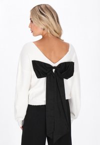 Weißer Pullover mit tiefem V-Ausschnitt hinten, der mit einer großen schwarzen Schleife verziert ist. Der Stoff sieht weich aus und hat eine gerippte Textur am Saum.
