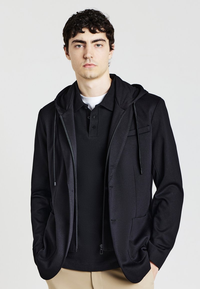 Schwarze Zip-Hoodie mit weicher Textur, über einem schwarzen Poloshirt getragen. Verfügt über eine dezente Fronttasche und eine verstellbare Kapuze mit Kordelzug.