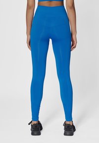 Högmidjade blå leggings med ett slätt, elastiskt tyg, med sid sömmar och en subtil texturdesign, tillsammans med svarta träningsskor.