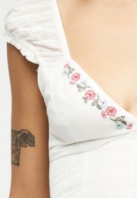 Λευκό φόρεμα με ανθισμένη κέντηση κοντά στον λαιμό και βολάν στα κοντά μανίκια, που δείχνει μερικό τατουάζ στο αριστερό μπράτσο.