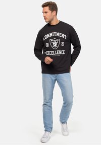 Schwarzes Langarm-T-Shirt mit weißem Text "ENGAGEMENT FÜR EXZELLENZ" und Raiders-Logo, kombiniert mit hellblauen Jeans und weißen Turnschuhen.