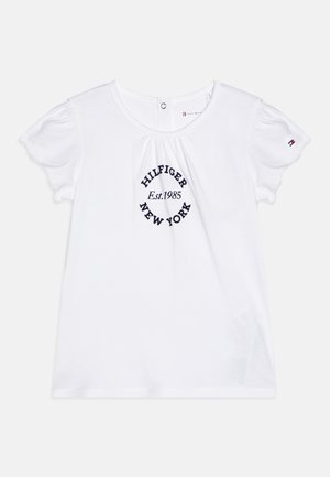 Hvit bomull t-skjorte med korte pufferm, med en marineblå sirkellogo som sier "HILFIGER Est.1985 NEW YORK." Knappelukking bak på nakken.