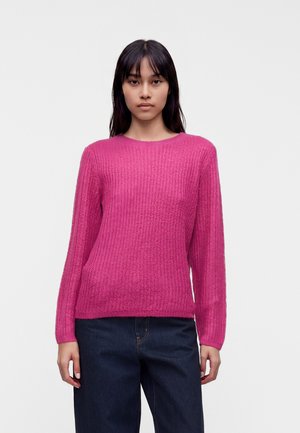 Kvinde med glat sort hår iført en knaldrød striktrøje og mørkeblå jeans, stående mod en ensfarvet lys baggrund.
